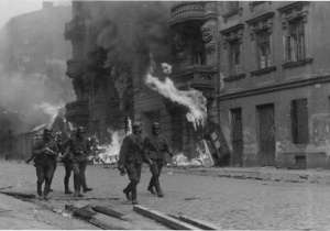 Grupa uzbroionych żołnierzy niemieckich idzie na tle ruin palącego się miasta